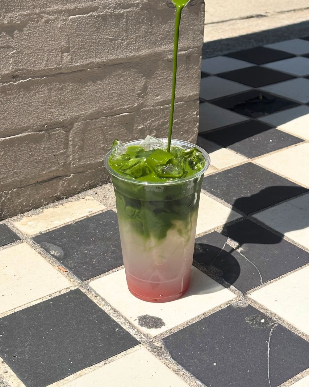 Matcha being poured into a strawberry milk matcha over ice, outside on the checkerboard tile